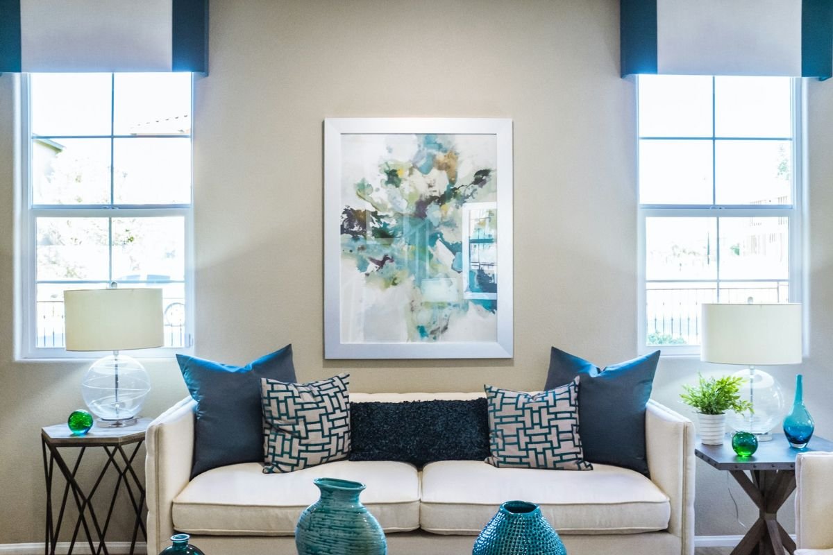 A stylish living room with a white sofa, abstract wall art, and blue accent pillows. There are two side tables with lamps and vases, and large windows on either side.