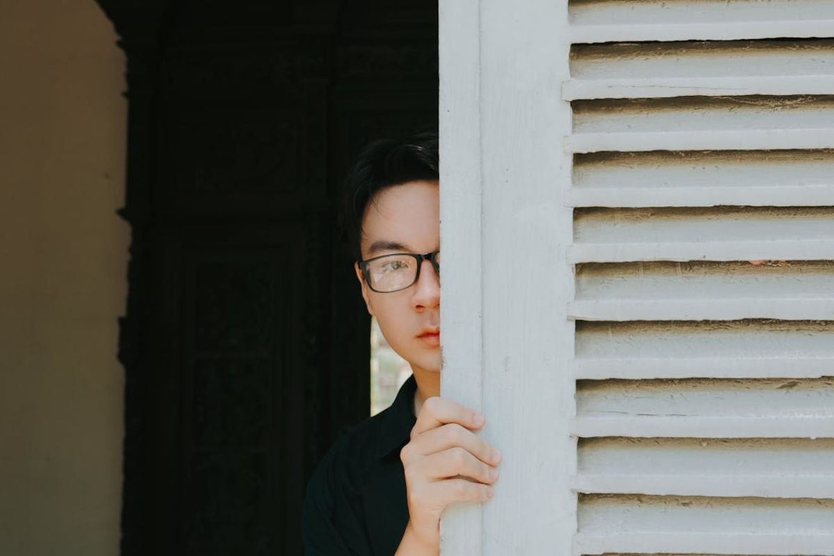 Avoiding Responsibility: The Art of Hiding Man peeking from behind a white louvered wall with a mischievous expression, suggesting he is avoiding something.