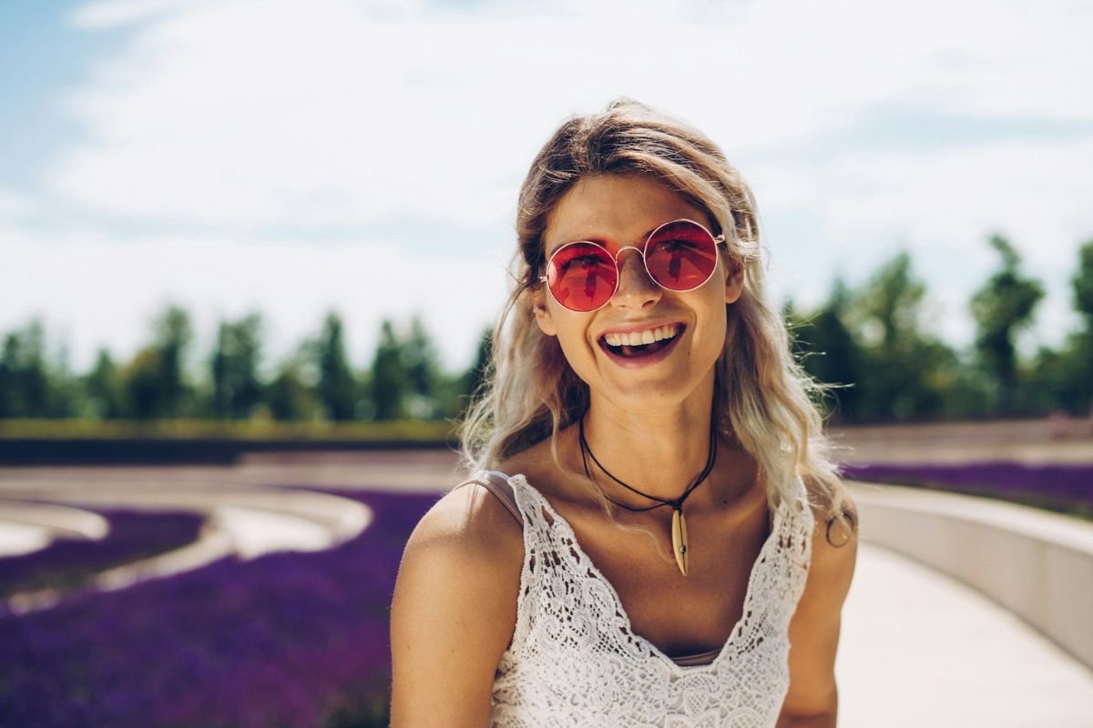 Woman Smiling in Red-Tinted Sunglasses A smiling woman with round red-tinted sunglasses, wearing a white lace top and a necklace, standing outdoors on a sunny day.