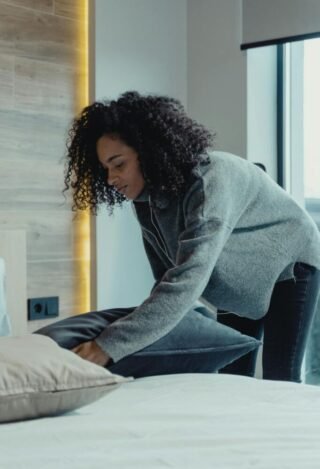 Woman casually placing down a pillow Woman in Gray Sweater and Blue Denim Jeans placing a pillow on a bed.