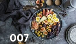 A colourful plate featuring figs, boiled eggs, grapes, cured meats, and a variety of cheeses with coffee mugs on the side.