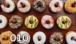 A top-down view of a wooden table covered with an assortment of colourful doughnuts. Each doughnut has a different topping, including chocolate, nuts, coconut flakes, and icing swirls. A small chocolate stick is placed in the centre of each doughnut.