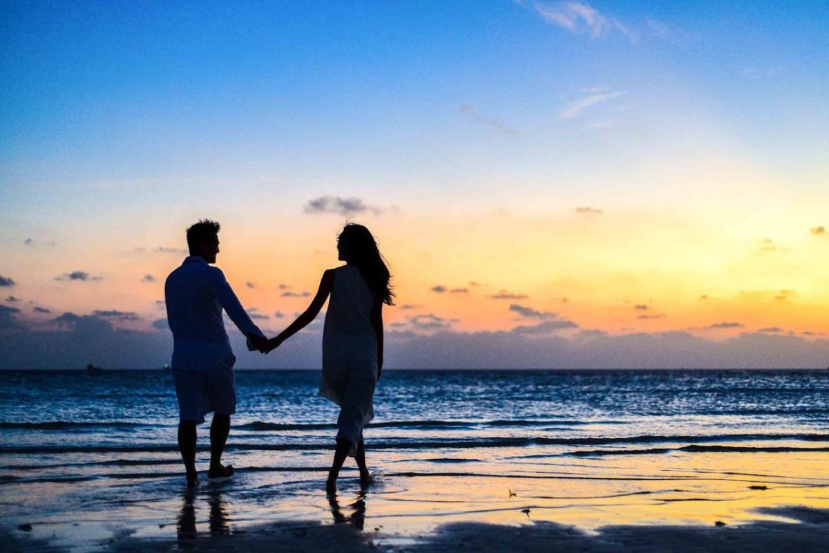 Couple Walking Hand in Hand on the Beach at Sunrise Man and woman holding hands while walking on the seashore during sunrise, silhouetted against the warm glow of the sun.