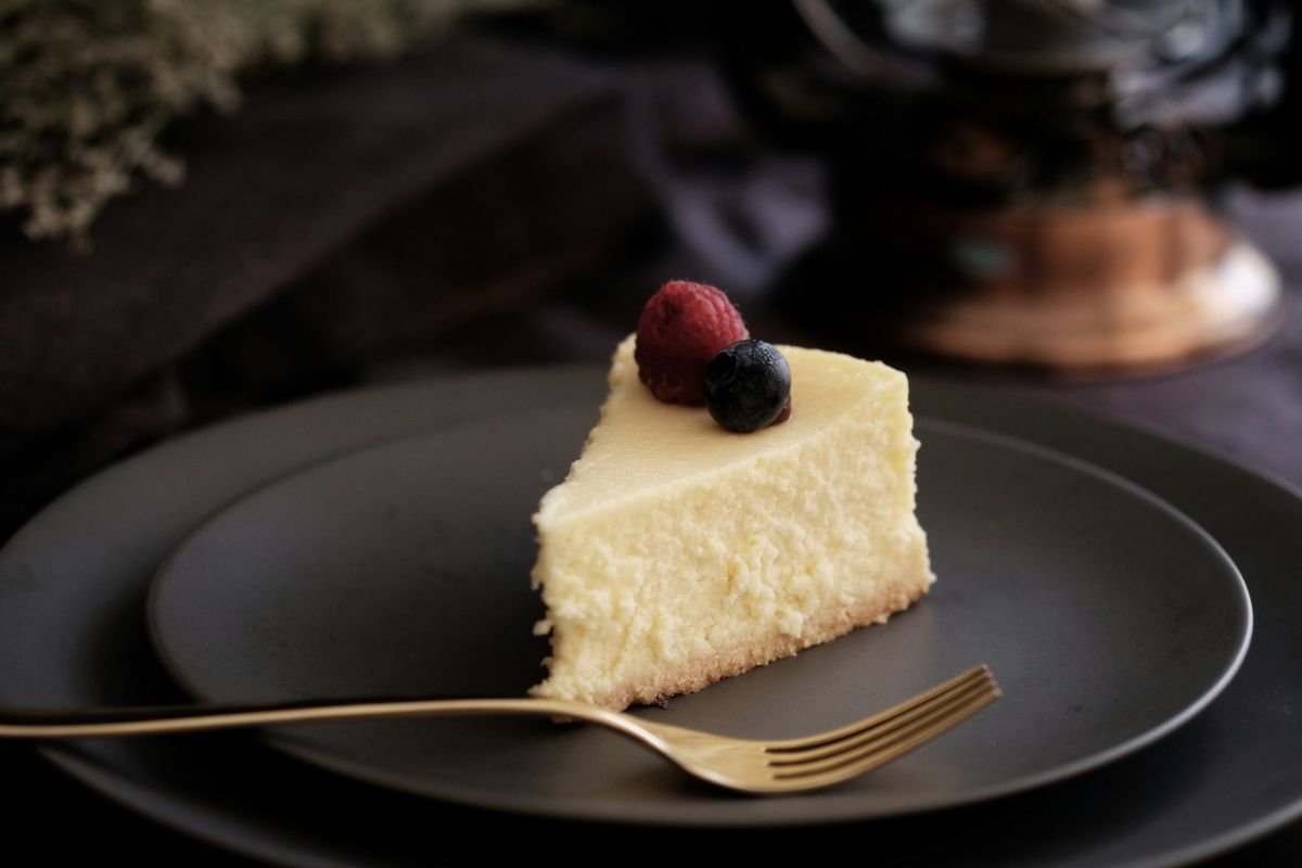 New York Cheesecake Slice A slice of creamy cheesecake garnished with a raspberry and a blueberry, served on a black plate with a gold fork.