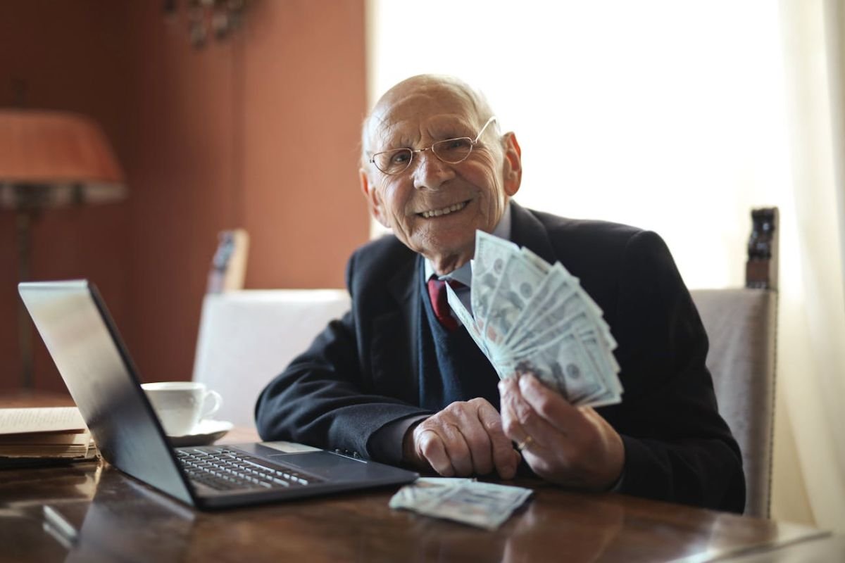 Managing Finances in Old Age: A Symbol of Clarity and Responsibility A cheerful senior businessman holding a stack of cash while working on a laptop, representing financial clarity and planning in later life.