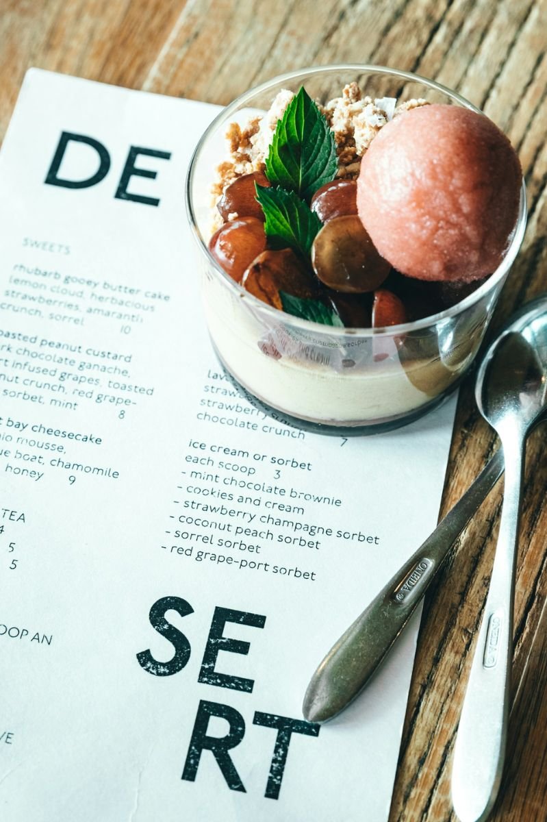 A glass of sorbet with grapes, mint garnish, and dessert crumbs placed next to a restaurant dessert menu on a wooden table.