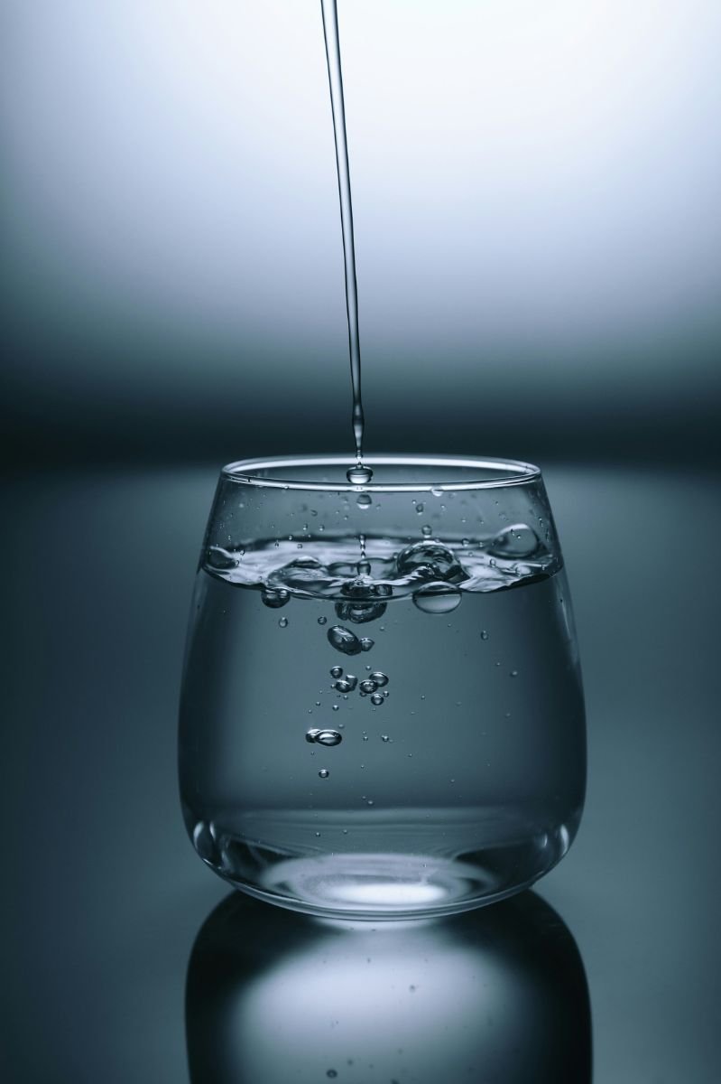 A stream of vinegar being poured into a transparent glass of water, creating ripples and bubbles as the liquid mixes, set against a cool, gradient background.