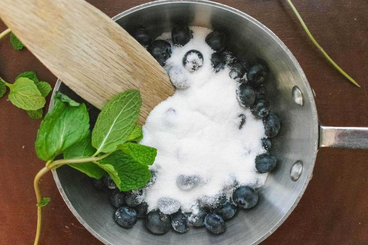 Blueberries and Sugar in a Saucepan A saucepan filled with fresh blueberries and a large amount of sugar, being stirred with a wooden spatula. A sprig of fresh mint is placed beside the mixture.
