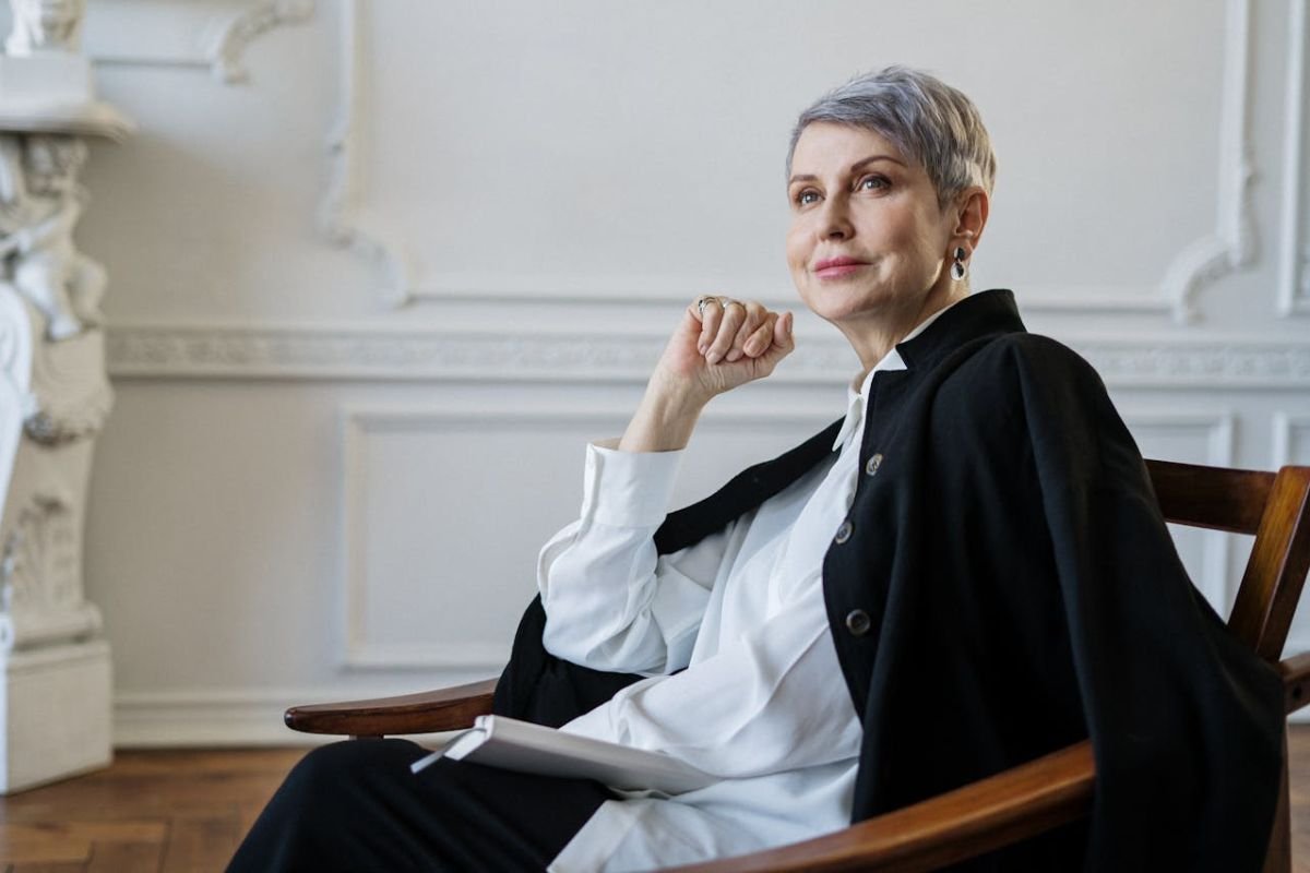 Confident Lady in Black Blazer Sitting Gracefully on a Chair A confident woman with short hair in a black blazer, sitting on a wooden chair in a composed manner.
