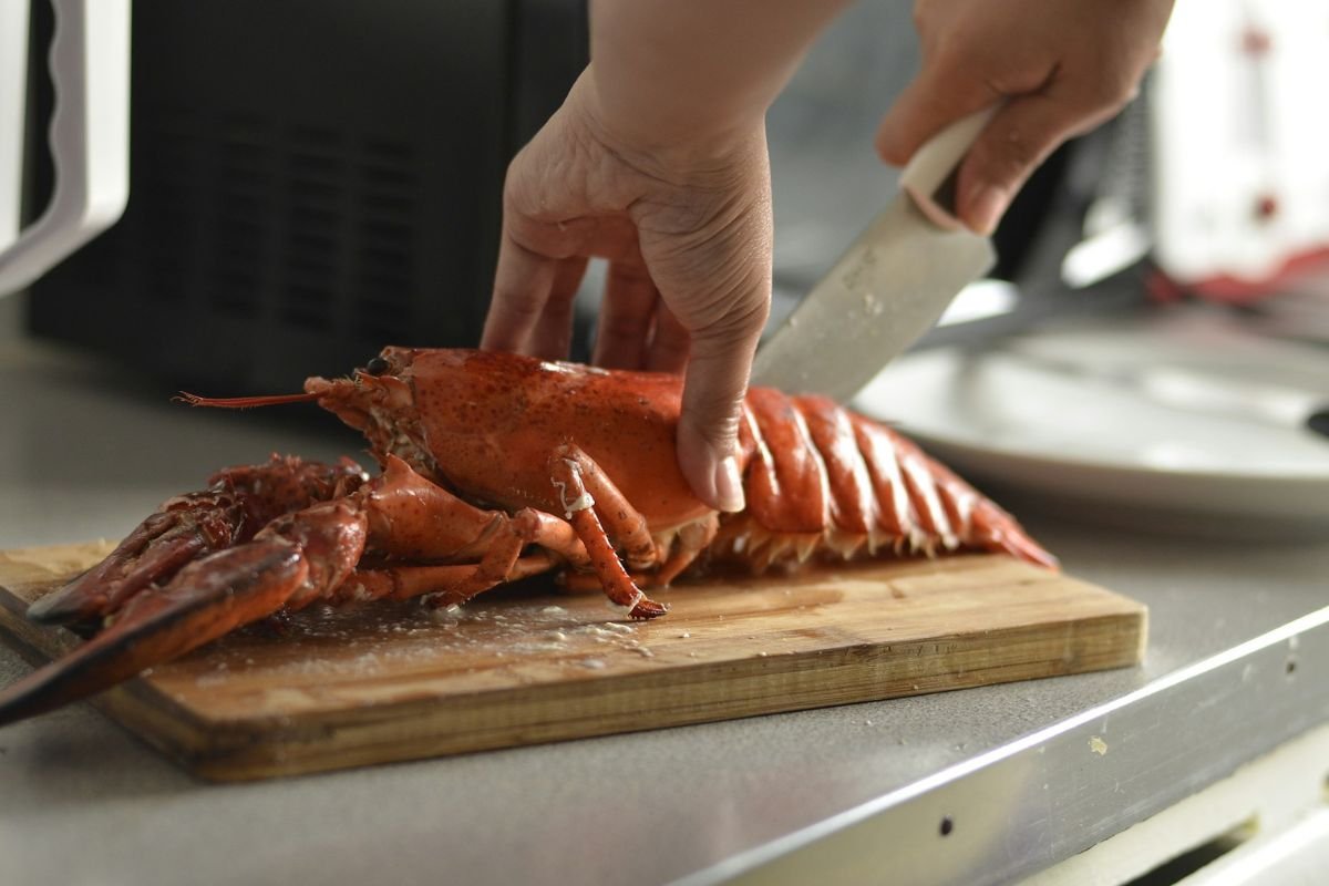 Cutting a Cooked Lobster in Half A person using a knife to cut a cooked lobster in half on a wooden cutting board.