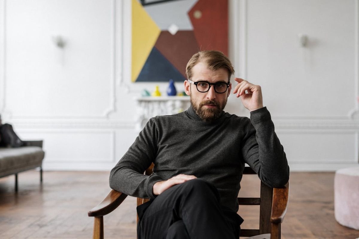 Thoughtful Man in Black Sweater Observing Intently A man in a black sweater sitting on a wooden chair, observing intently with a thoughtful expression.