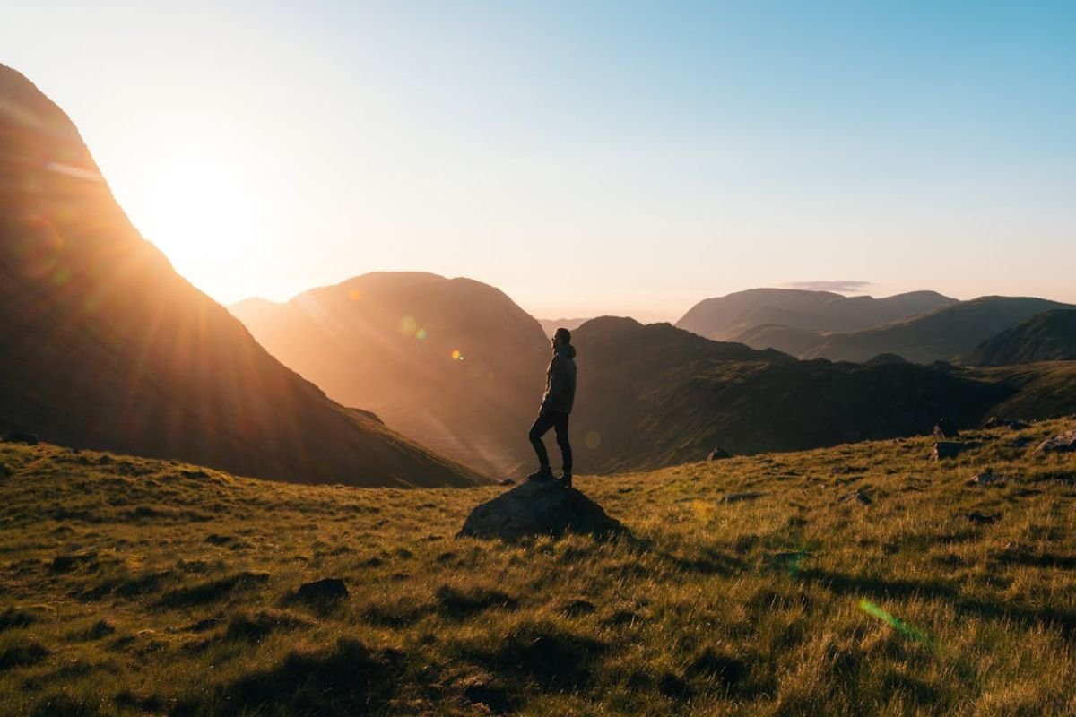 Reflecting on Life’s Three Key Relationships at Sunset A silhouette of a person standing on green grass with mountains in the background during golden hour, symbolising personal growth and reflection.