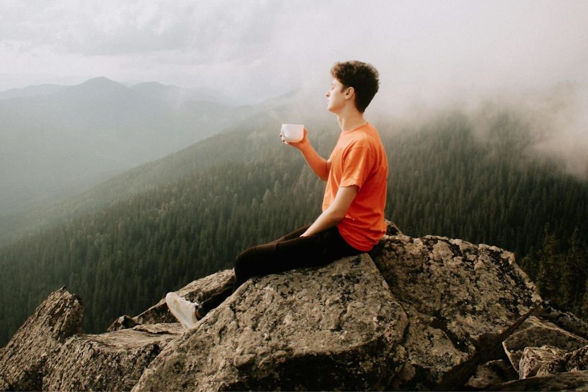 A man sitting on a rock in nature, holding a cup and enjoying the scenery, symbolising stepping away from commitments to live on one’s own terms.