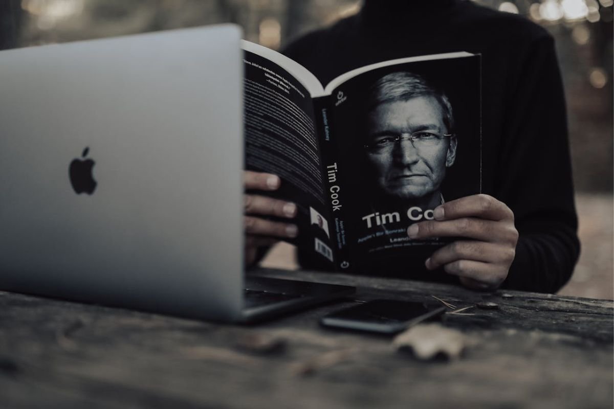 A person reading an autobiography while using a laptop, symbolising the power of daily reading for self-education, personal growth, and wealth-building habits.