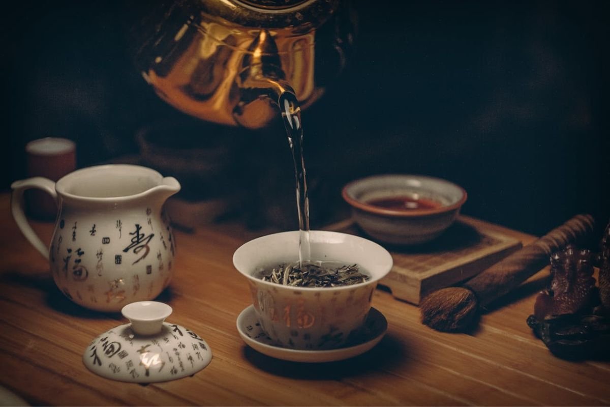 Gold Kettle Pouring Hot Water into a Ceramic Teacup A gold kettle pouring steaming hot water into a ceramic teacup on a wooden table, with a serene ambiance.