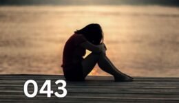 Sad woman sitting alone on a wooden deck, hugging her knees and looking out to sea, symbolising emotional pain and lack of self-love.