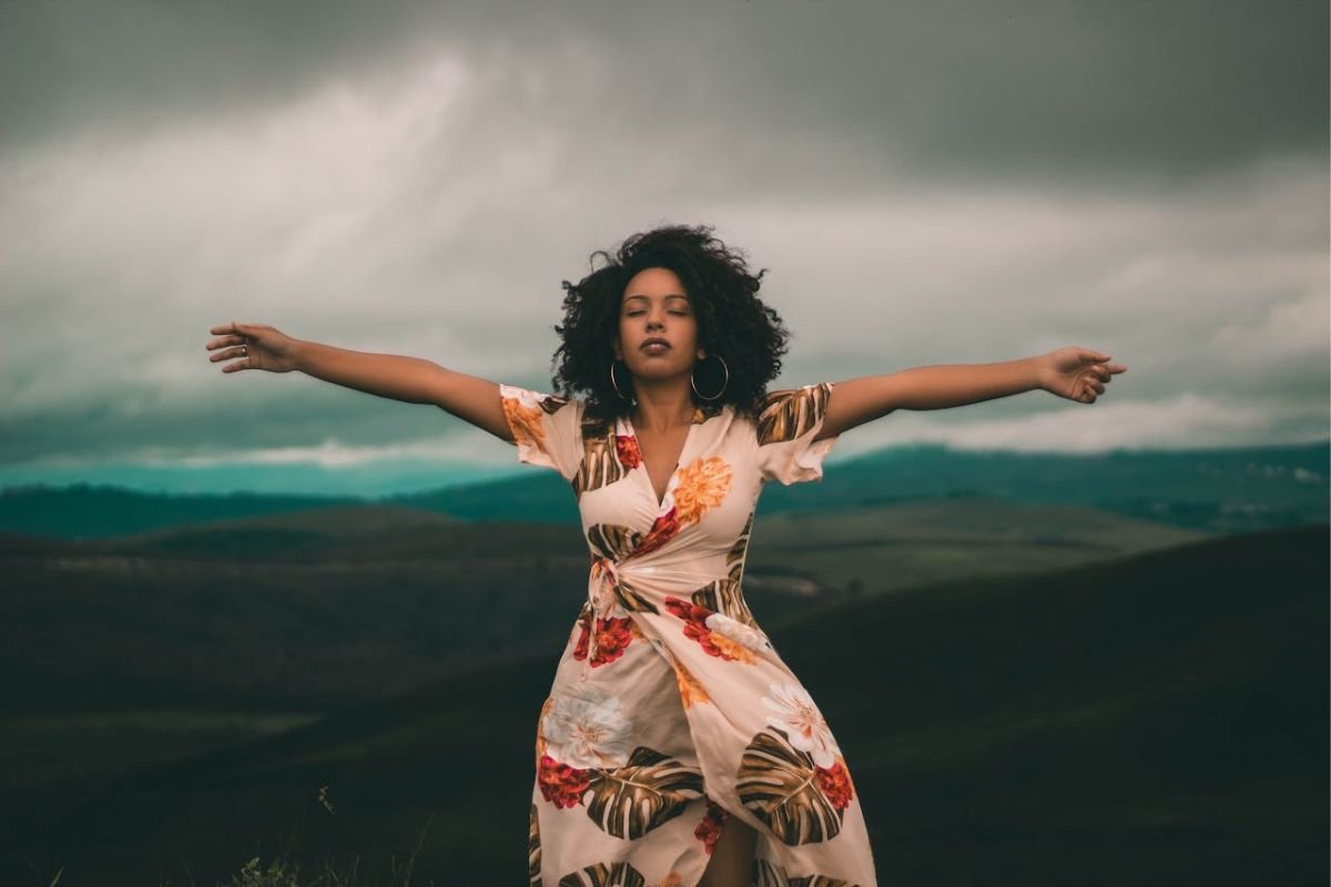 Woman Embracing Stillness in Nature: A Moment of Self-Reflection and Inner Listening Woman standing in a green grass field with eyes closed and arms gently extended, tuning into her emotions and listening inward.