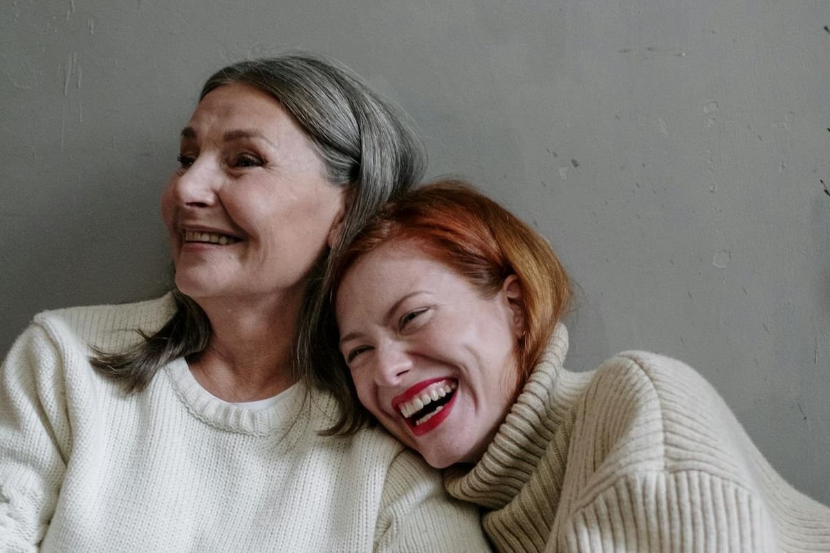 Elderly mother and adult daughter laughing together on a couch, showing a warm and joyful mother-daughter relationship in later life.