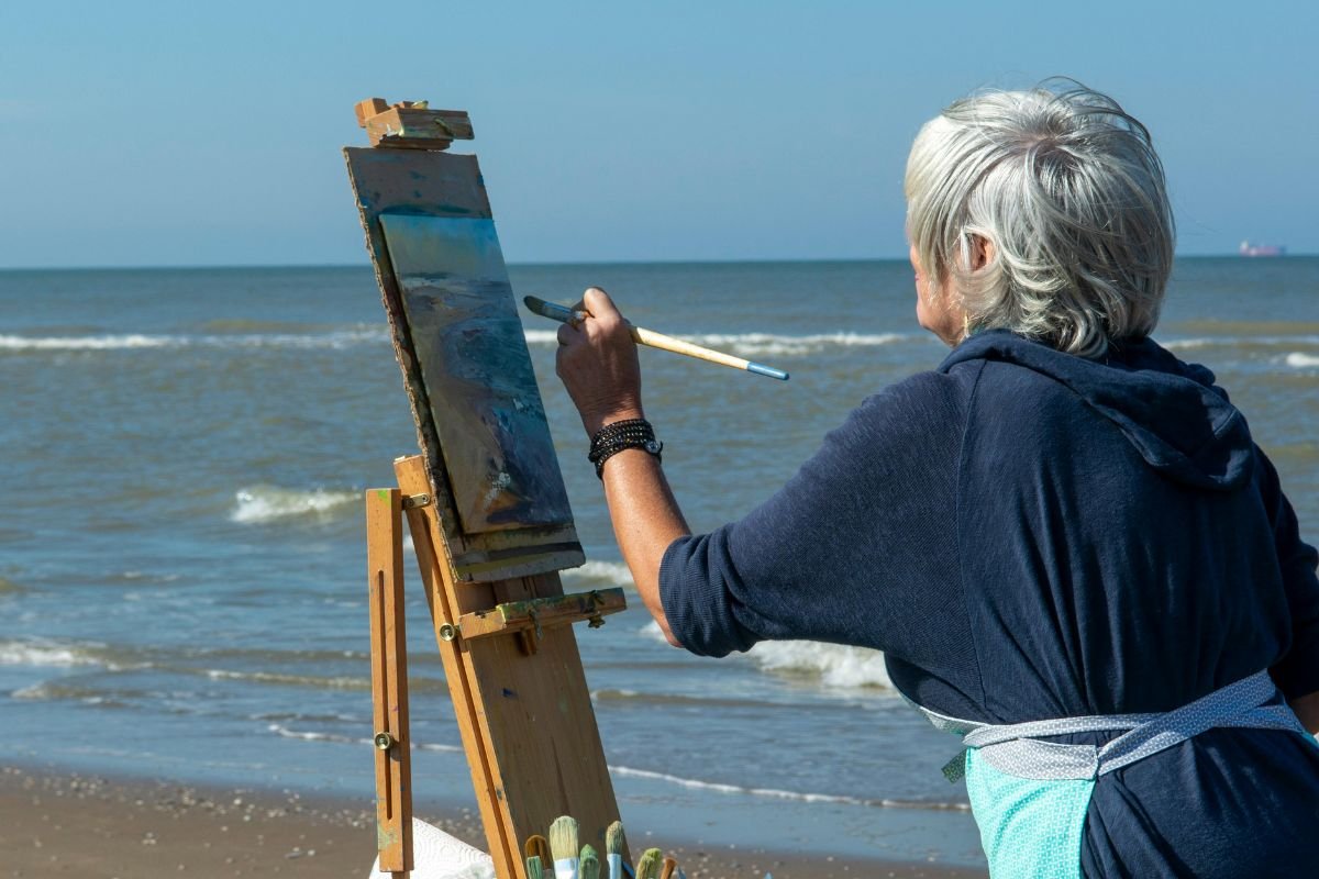 Elderly woman painting the beach where she stands, illustrating peaceful solitude, creativity, and self-discovery in later life.