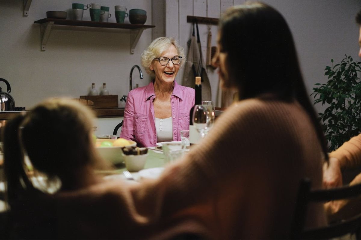 Elderly woman smiling and talking with family at the dinner table, illustrating uplifting, kind communication and family connection in later years.