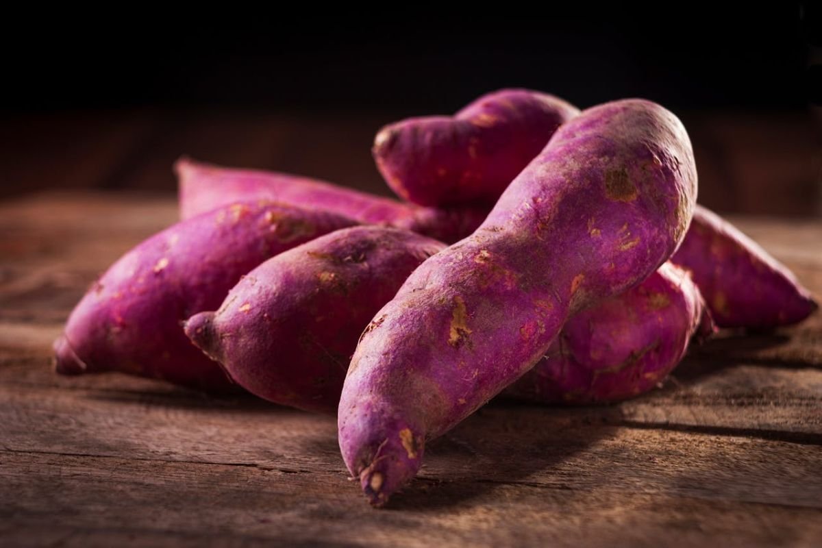 Vibrant Purple Sweet Potatoes: A Unique and Nutritious Variety A bunch of purple-skinned sweet potatoes resting on a rustic wooden surface, highlighting their rich colour and natural appeal.