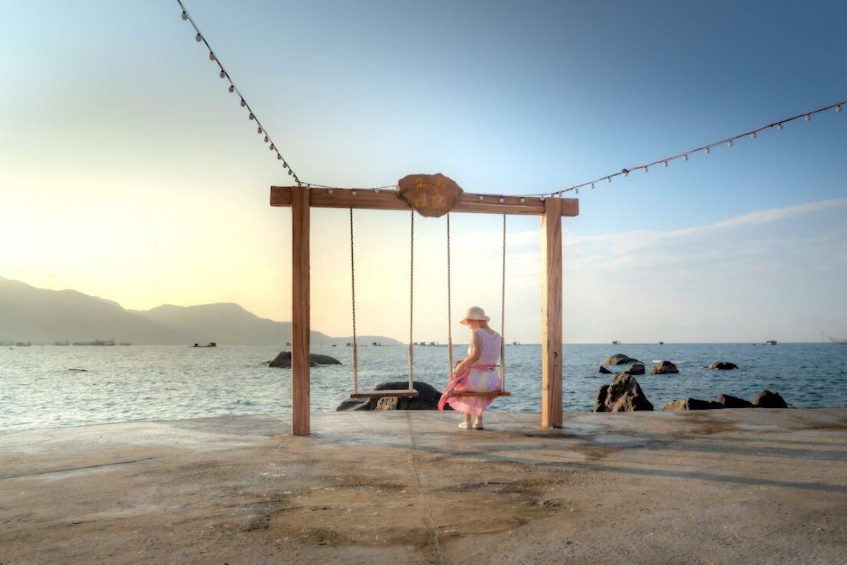 Elderly woman sitting on a swing by the sea, looking at an empty swing beside her, reflecting loneliness and aging.