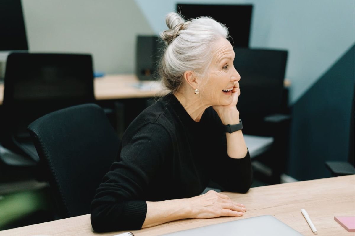Elderly woman smiling gently at her desk in a modern office, radiating warmth, humility, and mindful presence.