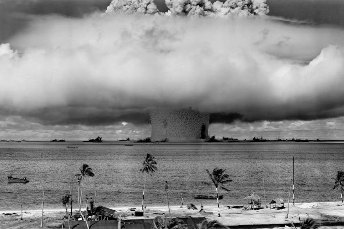 Grayscale photo of a massive explosion forming a mushroom cloud over the beach.