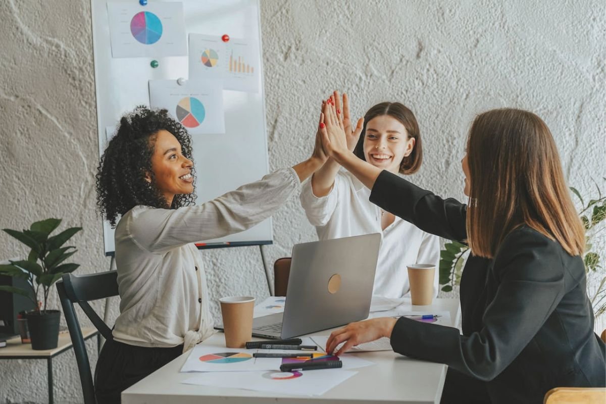 Group of women giving a high five at a work table, celebrating accomplishments and finding happiness through teamwork and meaningful work.