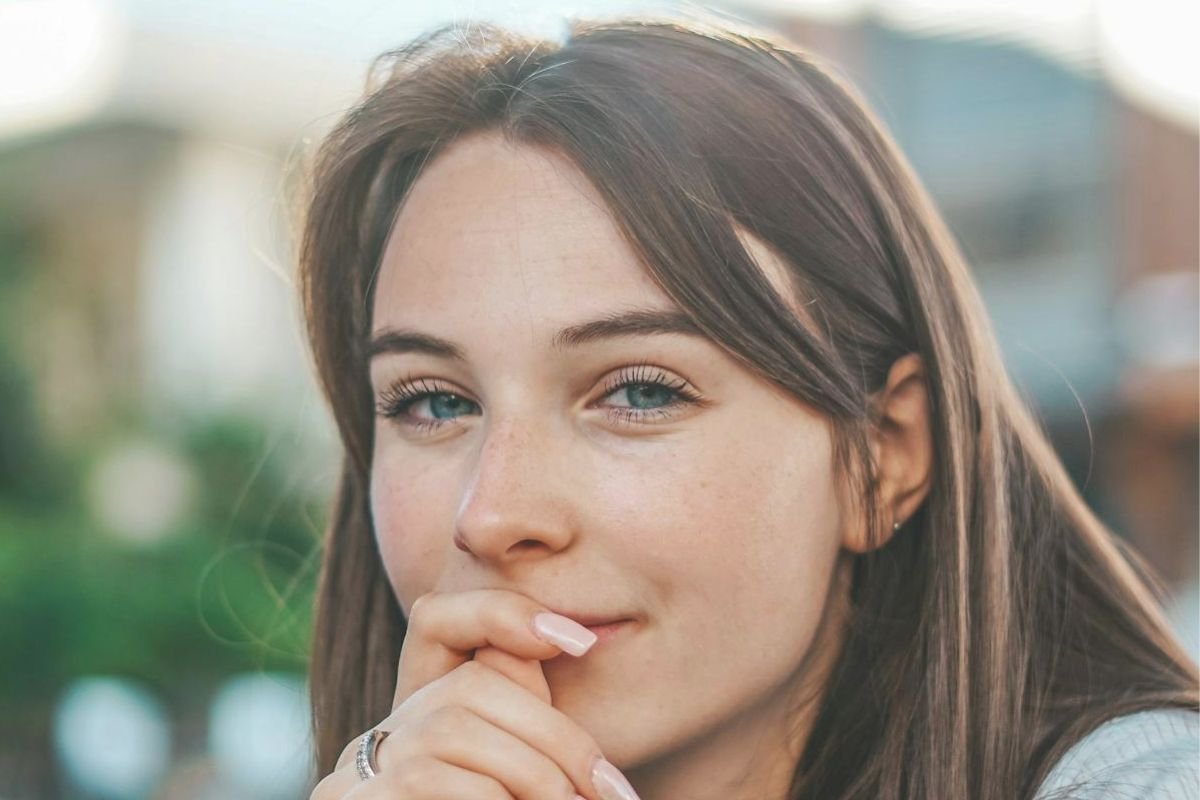 Woman, blue eyes, warm smile, hand near lips, conveying a moment of self-reflection