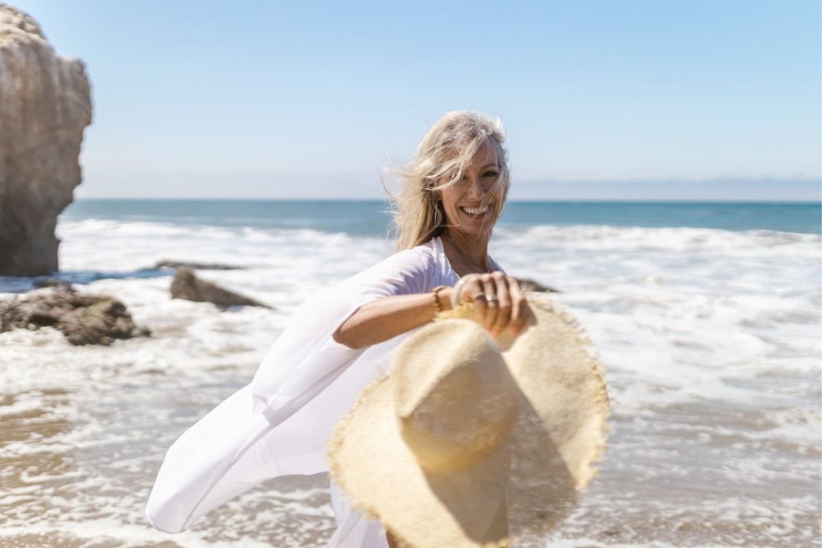 Mature woman smiling as she walks on the beach holding a straw hat, symbolising freedom, financial independence, and joy in later life.