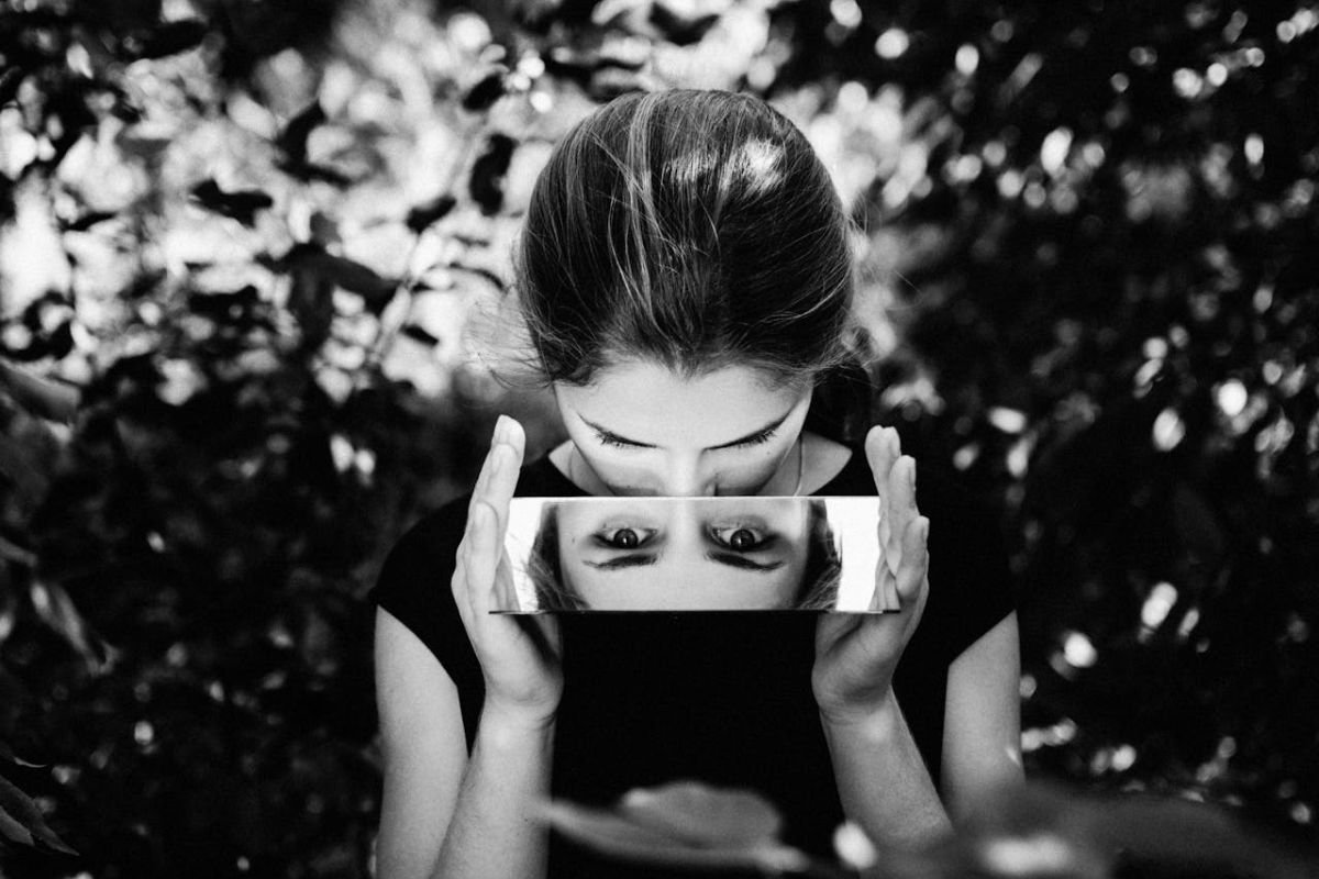 Black-and-white photo of a girl looking into a mirror, where her reflection shows a different emotion, symbolising Dissociative Identity Disorder (DID).