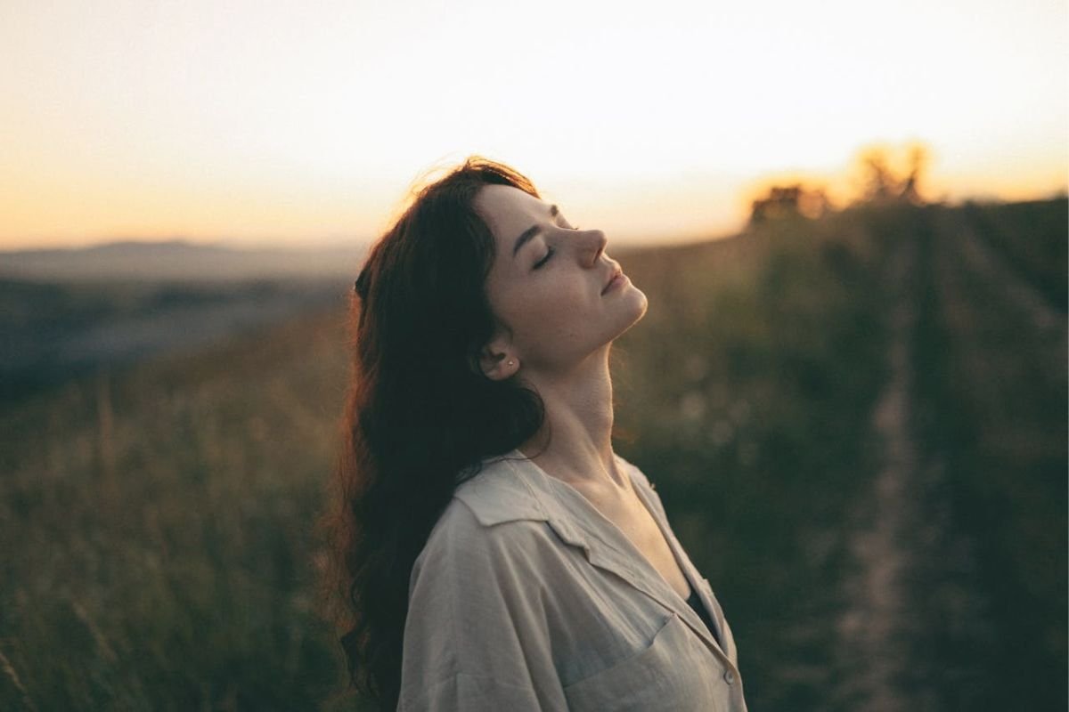 Woman standing in a meadow at sunrise with eyes closed, embracing mindfulness and calm after overcoming mental stress.