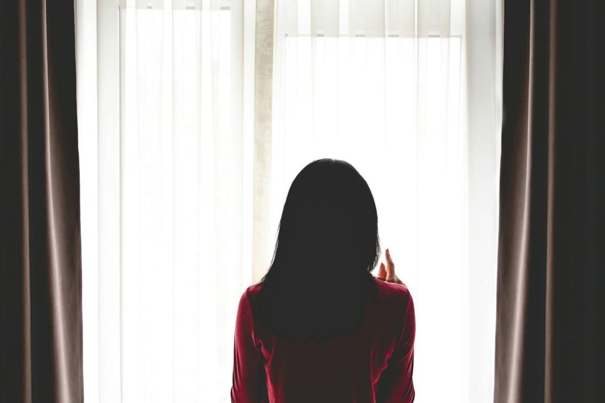 A woman standing by a window in soft shadows, symbolising self-reflection and the inner conflict behind using silent withdrawal in relationships.