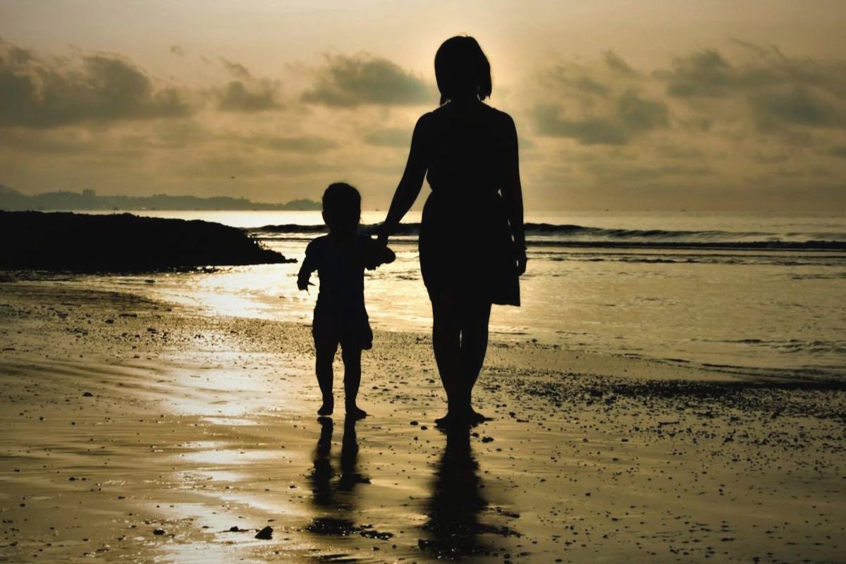 Silhouette of a mother holding her child’s hand walking on a beach, illustrating early attachment.