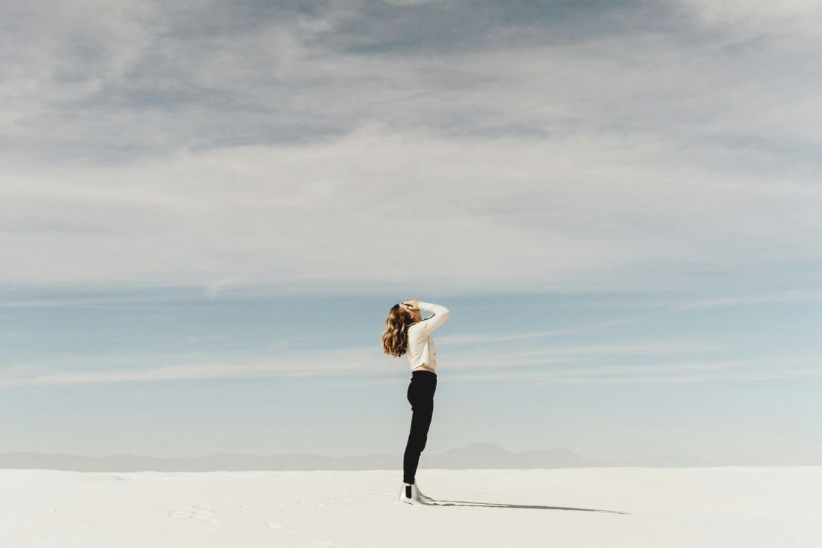 Woman standing on white sand with her face covered and head tilted upward, symbolising confusion, inner conflict, and uncertainty during periods of success.