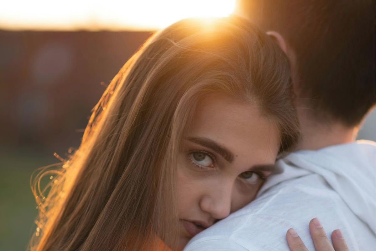 Woman hugging her partner at sunset with a distant expression, representing familiar but conflicted emotional attachment.