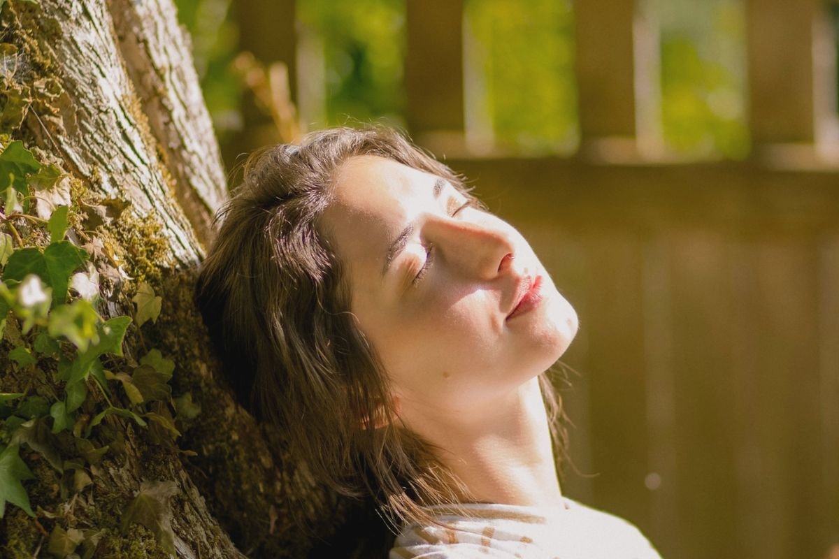 Woman lying against a tree in warm sunlight, representing modern lifestyles and the importance of natural sun exposure for healthy vitamin D levels.
