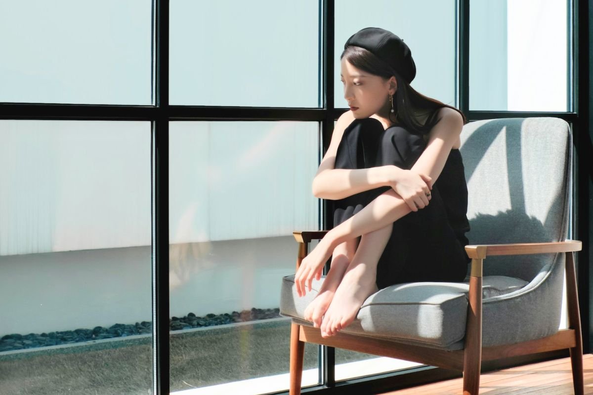 Woman sitting by a window looking outside, representing reflection, depression symptoms, emotional wellbeing, and mental health awareness.