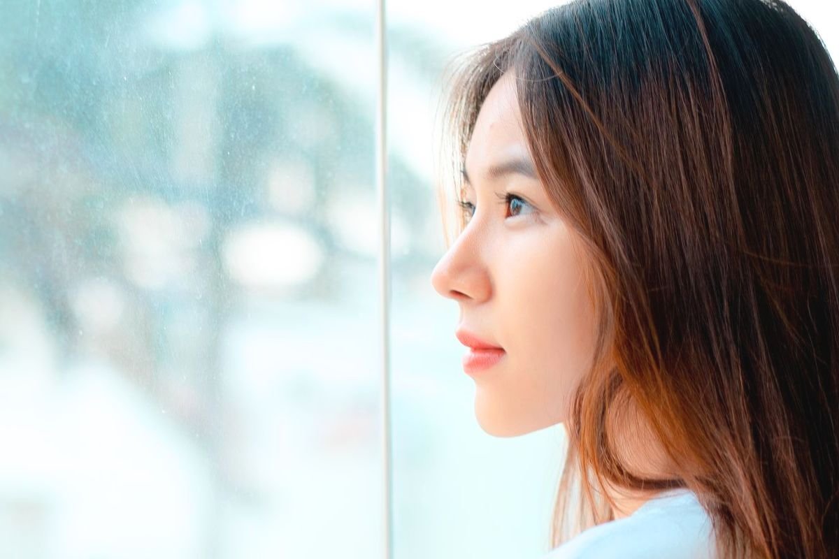 Young woman looking thoughtfully out of a window with a gentle smile, representing hope, self-compassion, and building confidence after social anxiety.